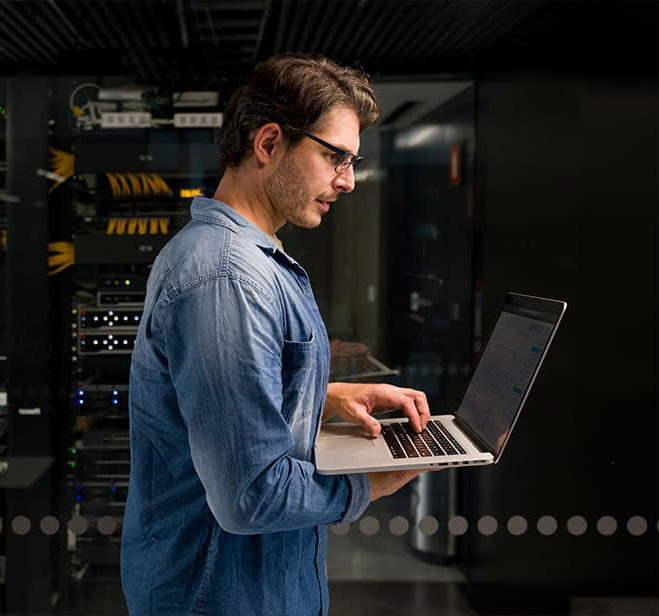 Male IT technician with glasses working on laptop in data center.