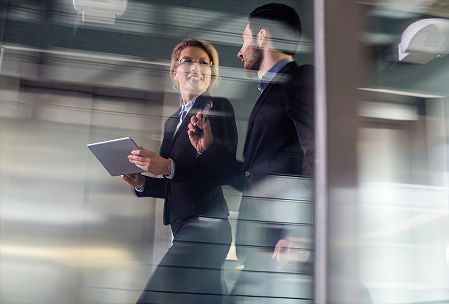 Male and female business professionals walking down hallway while referencing and discussing data displayed on a tablet.
