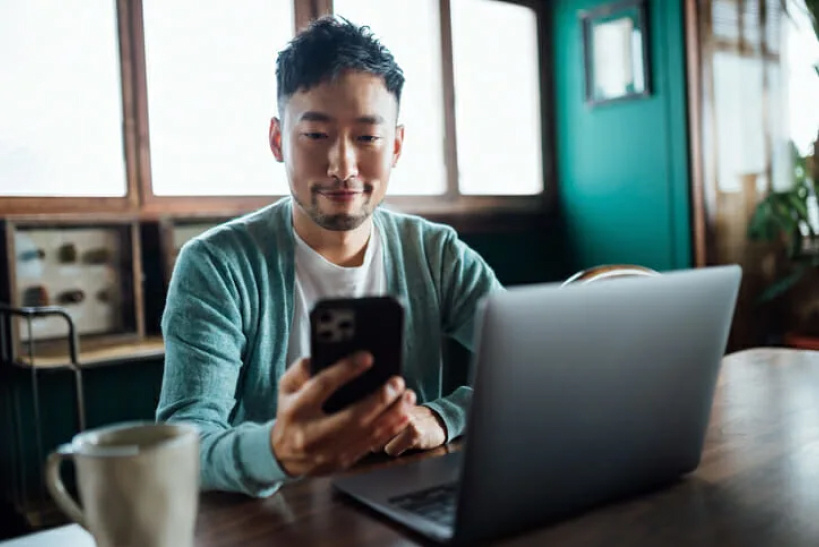 Young man in sweater accessing software from smartphone while working on laptop.