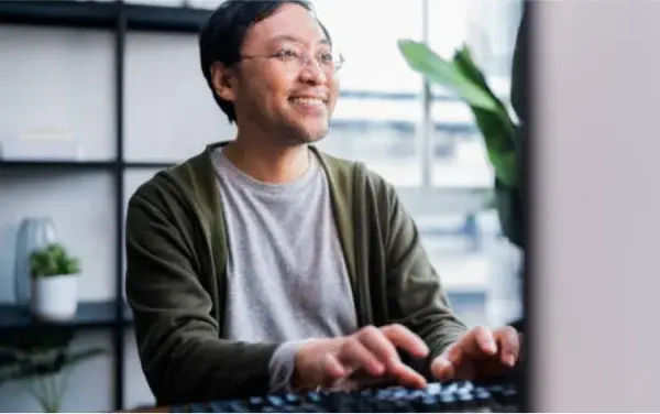Man in green sweater and glasses smiling while working at desktop computer.